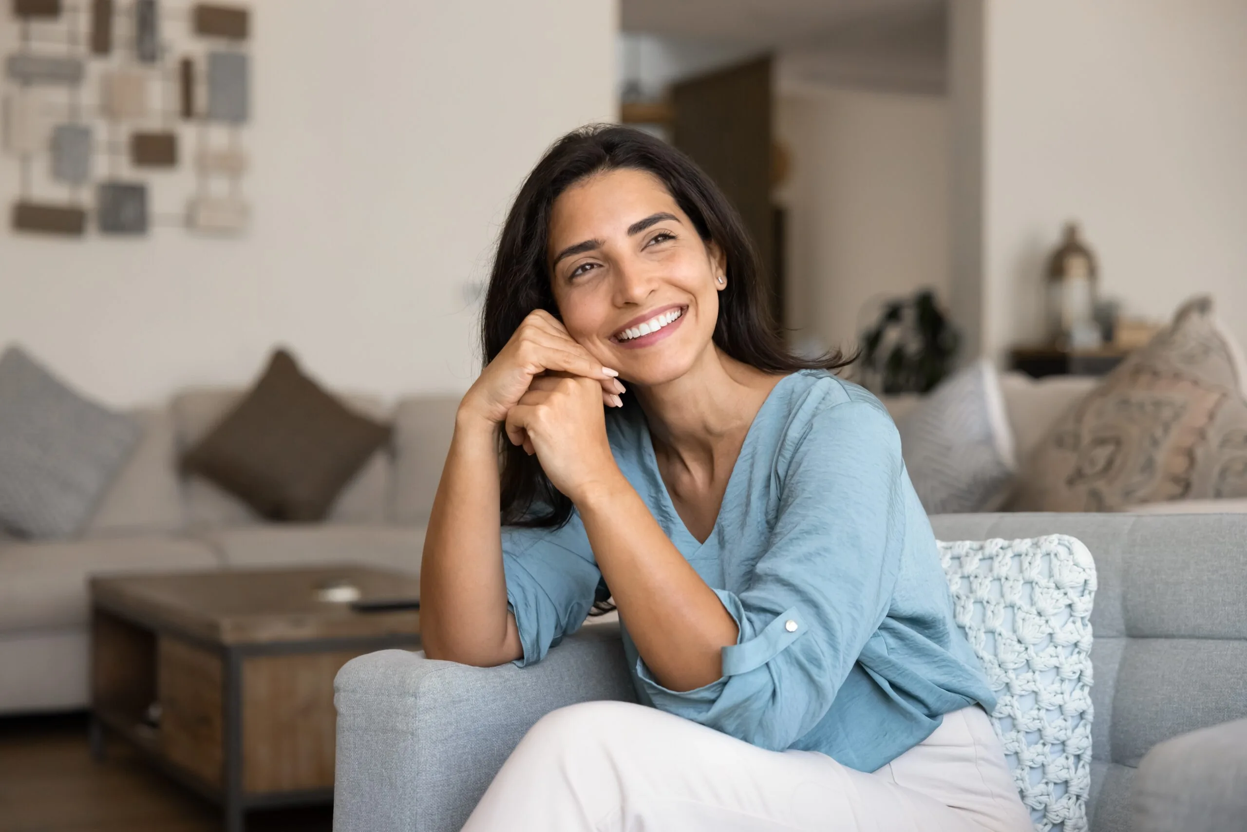 Cheerful beautiful young Hispanic woman sitting on comfortable couch, toothy smile