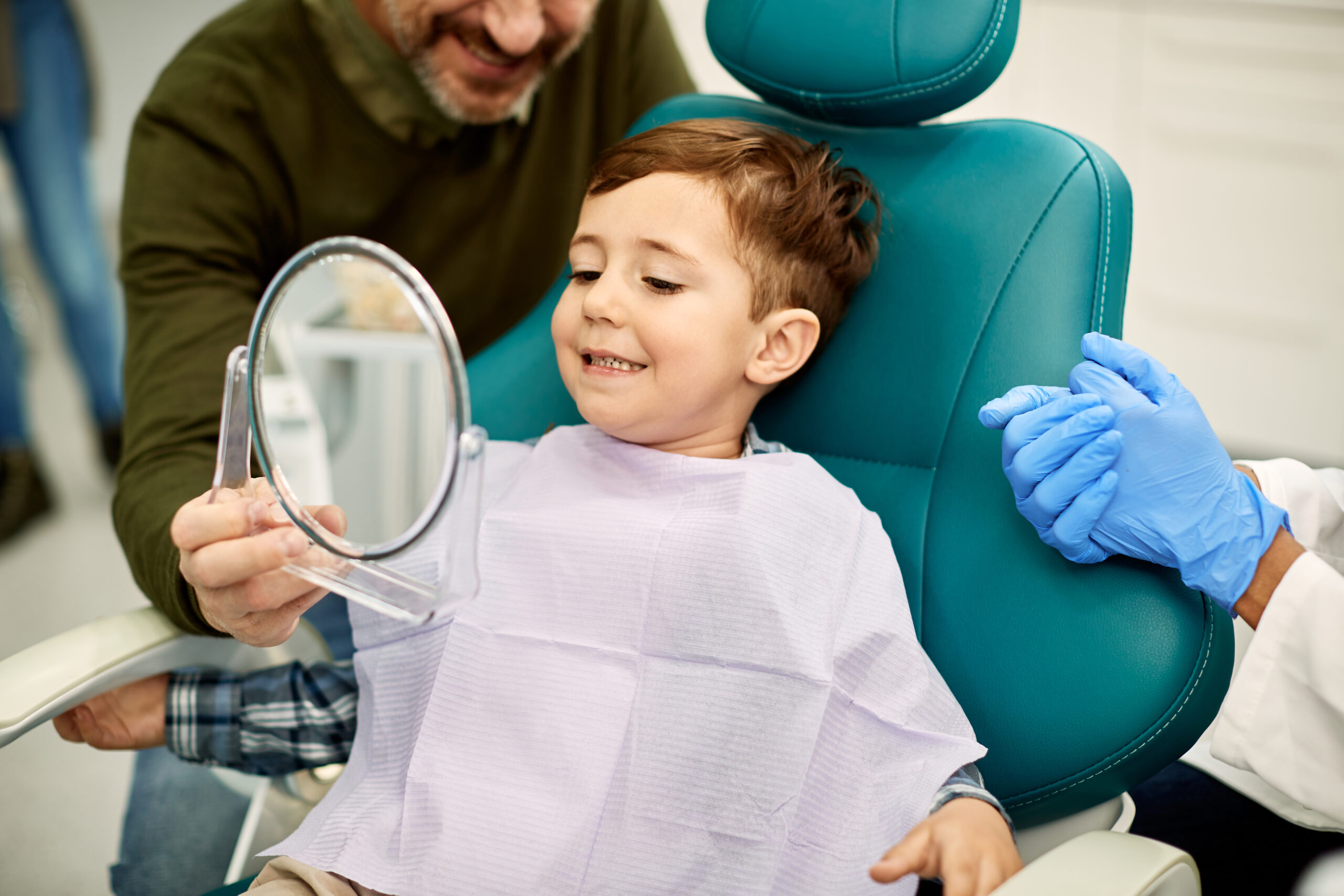smiling little boy holding up a mirror looking at teeth in dental office